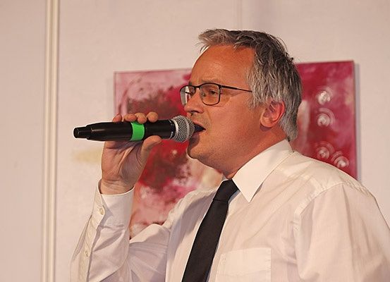 Ein Mann mit Brille singt in ein Mikrofon, während er ein weißes Hemd und eine schwarze Krawatte trägt. Er steht vor einem roten Gemälde.