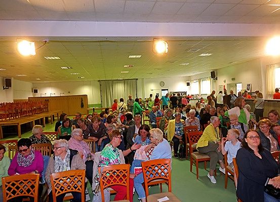 Eine große Gruppe von Menschen sitzt in Stühlen in einem Raum mit grünem Boden. Viele tragen Brillen und einige Halsketten. Eine Uhr ist an der Wand montiert.