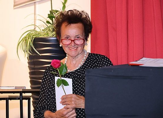 Eine ältere Frau in einem gepunkteten Kleid und mit Brille lächelt, hält eine rosa Rose und eine Karte. Hinter ihr steht ein Topf mit Pflanzen, ein Tisch mit Büchern und ein roter Vorhang.