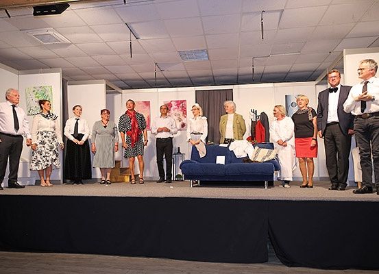 Eine Gruppe von Menschen auf der Bühne, einige feierlich gekleidet, stehen hinter einem blauen Sofa mit darauf liegenden Kleidern. Hinter ihnen hängen Kunstwerke an der Wand.