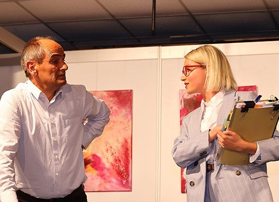 Ein Mann und eine Frau stehen in einem Innenbereich, möglicherweise einer Kunstgalerie. Der Mann sieht ernst aus, während die Frau eine Mappe in der Hand hält und zu sprechen scheint. Hinter ihnen hängen abstrakte Gemälde an der Wand.