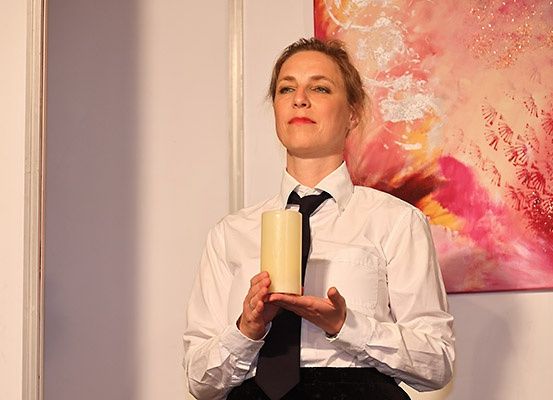 Eine Frau steht drinnen mit einer weißen Kerze, trägt ein weißes Hemd und eine schwarze Krawatte, mit einem farbenfrohen abstrakten Gemälde im Hintergrund.