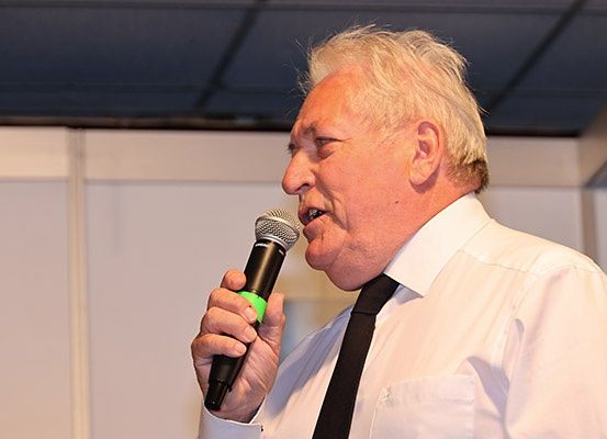 Ein älterer Mann spricht in ein Mikrofon. Er trägt ein weißes Hemd und eine schwarze Krawatte. Sein Haar ist kurz und blond.
