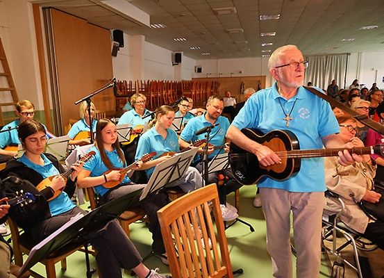Eine Gruppe von Musikern spielt in einem Raum, wobei ein älterer Mann vor einem Publikum Gitarre spielt.