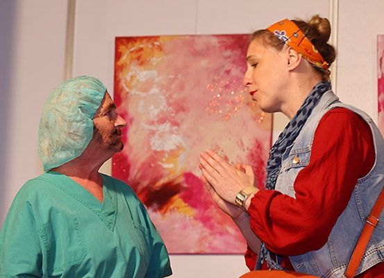 Ein älterer Mann in einem Krankenhauskittel und Mütze steht neben einer Frau mit einem Jeansweste, beide sehen ein abstraktes Gemälde an der Wand an.