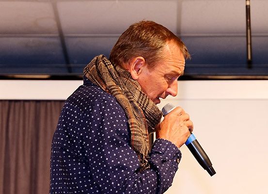 Ein Mann in einem blauen Punkthemd und einem karierten Schal hält ein Mikrofon und spricht oder singt. Er schaut leicht lächelnd nach unten. Hinter ihm befindet sich eine weiße Wand, ein brauner Vorhang und eine weiße Decke.