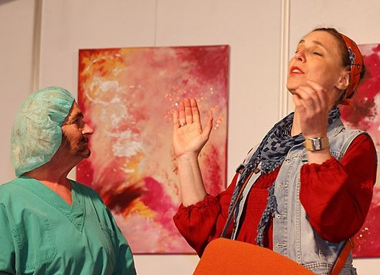 Ein Mann in grüner Arbeitskleidung und einem Haarnetz schaut eine Frau in einem roten Shirt an, die lächelt und ihre Hände vor ihr Gesicht hält.
