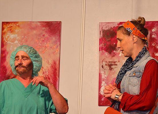 Ein Mann in grüner OP-Kleidung steht mit geschlossenen Augen und schaut eine Frau an, die eine Jeansweste und eine Uhr trägt. Sie stehen beide vor zwei abstrakten Gemälden.