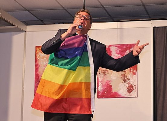 Ein Mann in Anzug und Brille tritt mit einer Regenbogenfahne um den Hals auf der Bühne auf. Er zeigt mit einer Hand nach links und hält in der anderen ein Mikrofon.