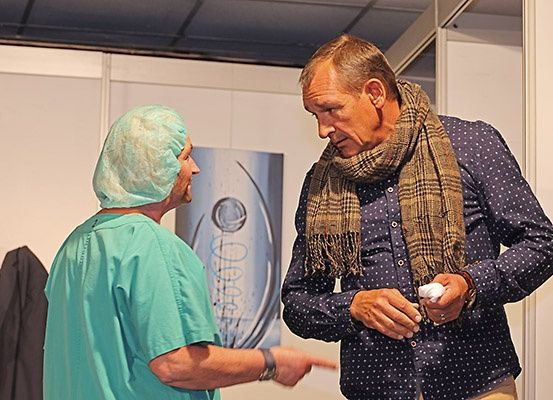 Ein älterer Mann in einem blauen Hemd und Schal spricht mit einem medizinischen Fachmann in grüner Arbeitskleidung. Eine weiße Wand mit einem silbernen Pokal ist im Hintergrund.