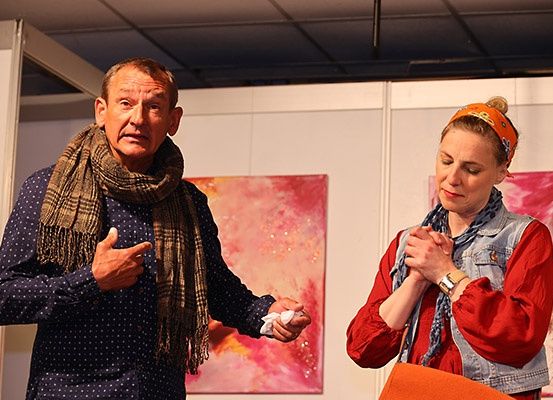 Ein älterer Mann und eine Frau stehen vor einer Wand mit zwei abstrakten Gemälden. Der Mann gestikuliert, während die Frau nach unten schaut, möglicherweise in einer Unterhaltung verwickelt.