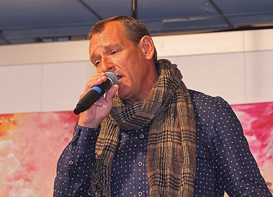 Ein Mann mit grauem Haar trägt einen karierten Schal und singt in ein Mikrofon in einem Raum mit einer weißen Wand und einem farbenfrohen Gemälde.