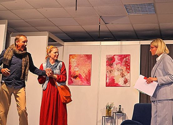 Drei Menschen stehen in einem Raum mit Gemälden an der Wand, einer hält Papiere und sieht ernst aus.