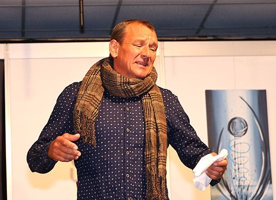 Ein Mann in einem blauen Punktehemd und einem karierten Schal steht in einem Raum und schaut nach oben, die Augen geschlossen. Er hält ein weißes Tuch in der rechten Hand. Hinter ihm ist eine weiße Wand und ein Banner mit goldenem Design.