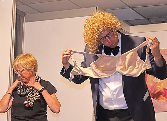 Ein Mann mit lockigem Haar und Fliege hält einen BH in den Händen, möglicherweise für ein Publikum. Eine ältere Frau steht in der Nähe und schaut ihn an.