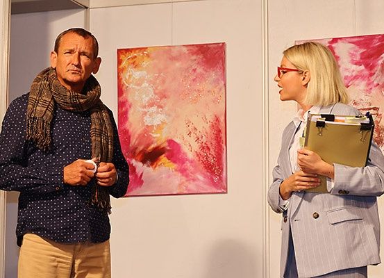 Ein Mann in einem Schal und eine Frau mit Brille stehen vor zwei abstrakten Gemälden.