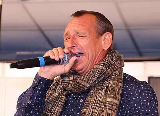 Ein Mann mit geschlossenen Augen singt in ein Mikrofon, trägt einen Schal und ein blaues Hemd mit weißen Sternen.