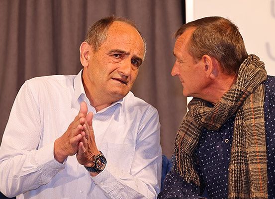 Zwei Männer sitzen und unterhalten sich in einem Konferenzraum. Der Mann auf der linken Seite klatscht. Der Mann auf der rechten Seite trägt einen karierten Schal.