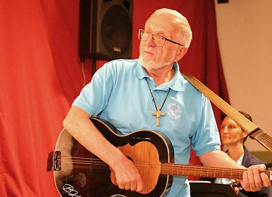 Ein älterer Mann in einem blauen Shirt spielt eine Gitarre, trägt eine Brille und eine Kreuzkette. Eine Frau steht hinter ihm und hält eine weitere Gitarre. Ein Lautsprecher ist an einer roten Wand montiert.