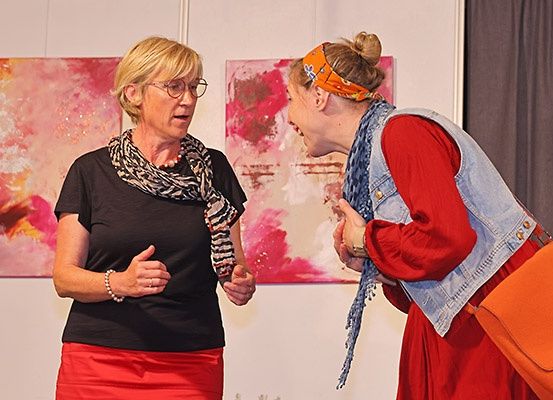 Zwei Frauen stehen vor einer Wand mit leuchtenden Gemälden, eine in einer Jeansweste und einer orangefarbenen Tasche, die andere in einem schwarzen Shirt und einem roten Rock, in einer Unterhaltung verwickelt.