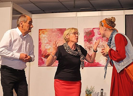 Drei Menschen stehen in einem Raum mit abstrakten Gemälden an der Wand. Ein Mann trägt ein weißes Hemd und schwarze Hose, während zwei Frauen schwarze und rote Kleider tragen.