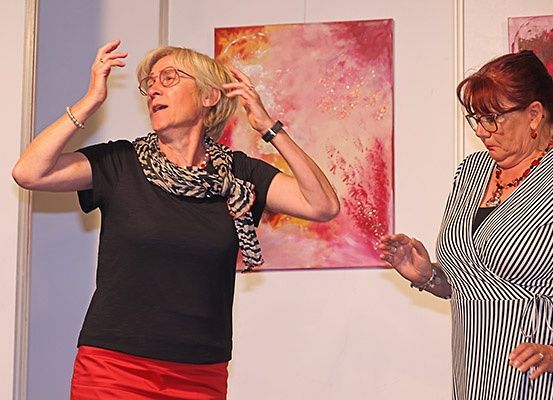 Zwei Frauen sind drinnen. Eine gestikuliert mit ihren Händen, während die andere zusieht. Sie stehen vor einem rosafarbenen Gemälde.