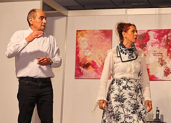 Ein älterer Mann und eine Frau stehen in einer Kunstgalerie, der Mann gestikuliert, während die Frau nach vorne schaut. Hinter ihnen zieren zwei große abstrakte Gemälde die Wand.
