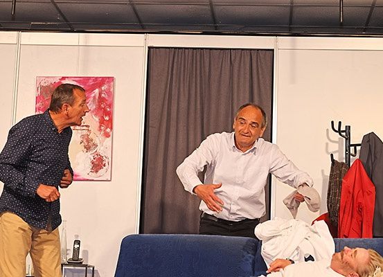 Drei Männer stehen und unterhalten sich in einem Innenbereich. Ein Mann gestikuliert mit den Händen, ein anderer lächelt und schaut den Mann an, der gestikuliert. Ein dritter Mann liegt mit einem weißen Bademantel auf einer blauen Couch. Ein roter Mantel hängt hinter ihnen an einem Kleiderständer.