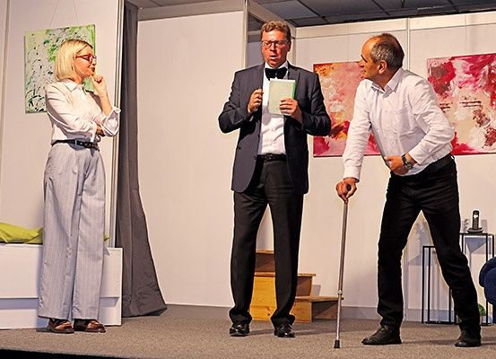 Drei Personen stehen auf einer Bühne, ein Mann hält ein Buch und einen Stock, ein anderer Mann schaut in das Buch, und eine Frau steht neben einem Bett mit einem Kissen.