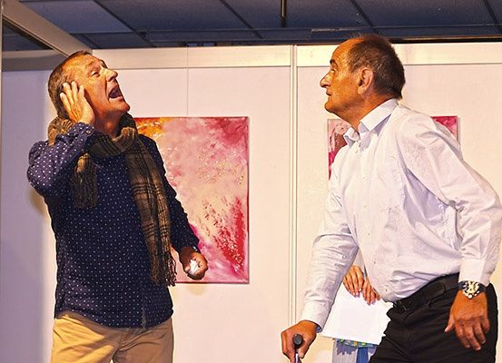 Zwei Männer stehen in einer Kunstgalerie. Einer wirkt schockiert und hält seine Hände an die Ohren, während der andere mit einem Stock schaut. Kunstwerke hängen an der Wand hinter ihnen.