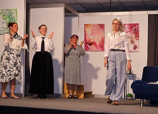 Fünf Frauen in formeller Kleidung stehen auf der Bühne und klatschen. Drei sind links, eine in der Mitte und eine rechts. Hinter ihnen befinden sich Gemälde an der Wand, eine Couch und ein Tisch.