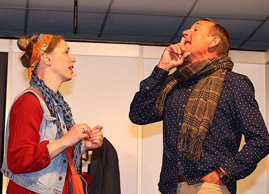 Ein Mann und eine Frau stehen drinnen und sehen sich an. Die Frau trägt eine Jeansweste und einen Schal. Der Mann ist in einem blauen Hemd mit einem Schal um den Hals gekleidet und sieht überrascht aus.