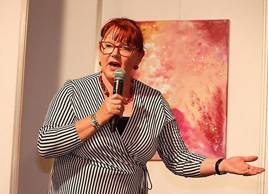 Eine Frau mit roten Haaren, die eine Brille, eine Halskette und ein gestreiftes Kleid trägt, hält ein Mikrofon. Sie scheint in dieses zu sprechen. Ein abstraktes Gemälde mit roten und pinken Farben befindet sich hinter ihr.