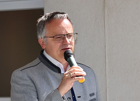 Ein Mann mit Brille und grauer Jacke spricht in ein Mikrofon und steht vor einer beigen Wand.