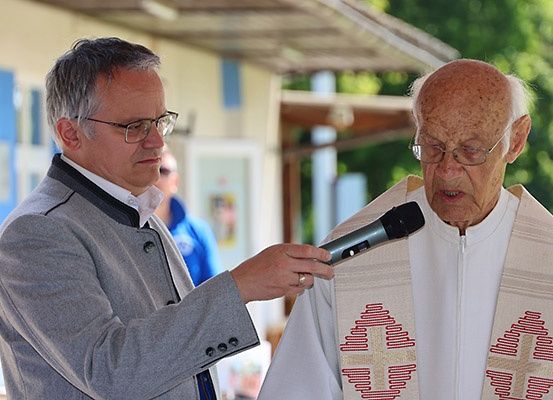 Ein älterer Priester mit einer weißen Robe mit roten Kreuzen und einem älteren Mann mit Brille, der ein Mikrofon hält.