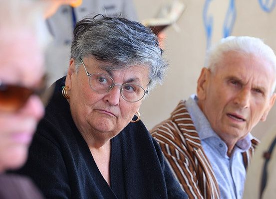 Drei ältere Personen, eine Frau mit Brille und ernster Miene, ein Mann in einem gestreiften Hemd und eine weitere Person mit Sonnenbrille, sitzen gemeinsam in einem Raum.
