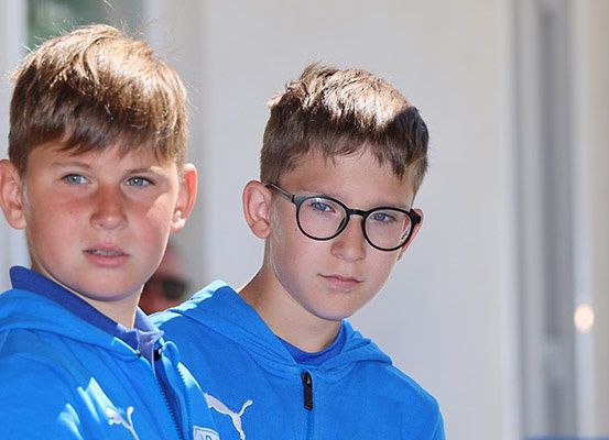 Zwei junge Jungen in blauen Puma-Kapuzenpullovern, einer mit Brille, stehen zusammen in einem hellen, gut beleuchteten Raum.