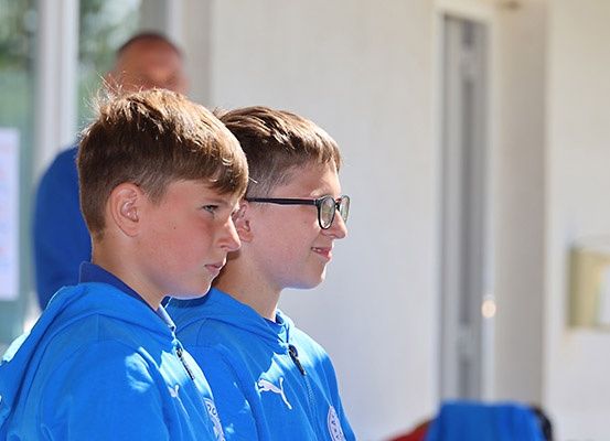 Zwei Jungen in blauen Hoodies stehen nebeneinander, einer trägt eine Brille. Hinter ihnen ist eine verschwommene Figur im Hintergrund.