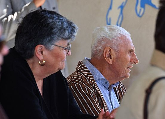 Ein älterer Mann und eine ältere Frau sitzen nebeneinander, nach rechts schauend, mit einer Person auf der linken Seite. Sie befinden sich in einer Innenumgebung mit einer Wand hinter ihnen.