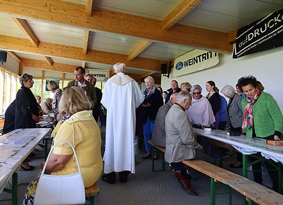 Eine Gruppe von Menschen versammelt sich in einem Saal. Einige sitzen auf Bänken, während andere stehen. Eine Person in einer weißen Robe steht vor ihnen. Werbebanner hängen an der Wand.