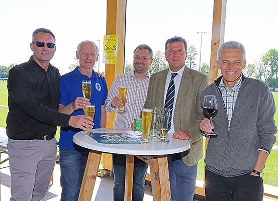 Fünf Männer stehen vor einem Tisch mit verschiedenen Getränken. Sie scheinen für ein Foto zu lächeln. Hinter ihnen ist ein Rasenfeld.