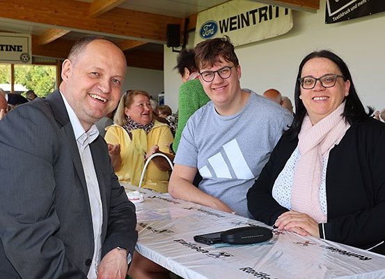 Drei Personen sitzen an einem Tisch und lächeln für ein Foto. Sie befinden sich in einer Halle mit einem Tisch, der mit einem weißen Tuch bedeckt ist. Hinter ihnen ist ein Banner mit dem Wort Ford zu sehen.
