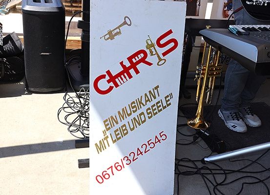 Ein weißes Schild mit roten Buchstaben und Trompeten. Es steht 'CHRIS' und die Telefonnummer 067613242545. Eine Trompete und ein Keyboard sind rechts.