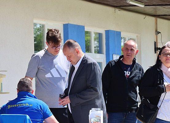 Fünf Menschen stehen vor einem Gebäude mit blauen Fensterläden. Ein Mann schaut nach unten, ein anderer trägt eine Brille und eine Frau trägt eine schwarze Tasche. Ein Mann in einem blauen Shirt sitzt auf einem Stuhl.