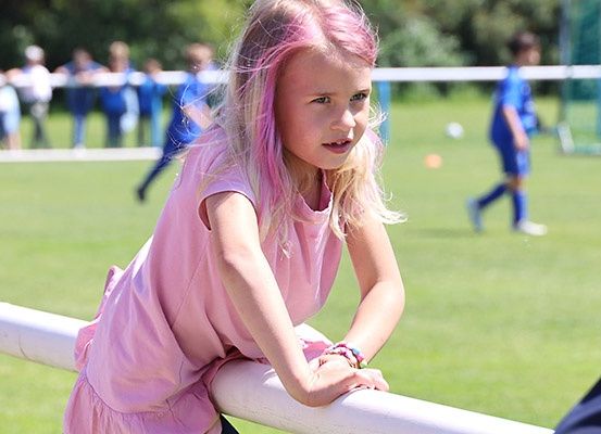 Ein kleines Mädchen mit pinken Haaren beobachtet ein Fußballspiel und steht auf einer weißen Brüstung. Im Hintergrund sind mehrere Spieler in blauen Trikots verschwommen.