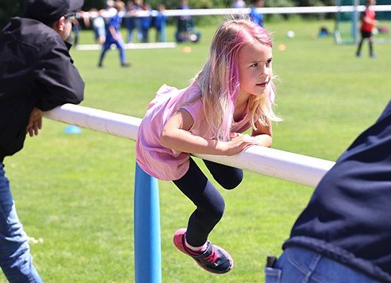 Ein junges Mädchen mit rosa Haaren klettert auf einen weißen Pfosten auf einem Rasenfeld. Sie trägt ein rosa Shirt und schwarze Hose. Hinter ihr steht eine weitere Person. In der Ferne spielen Menschen Fußball, und einige stehen in der Nähe des Tors.
