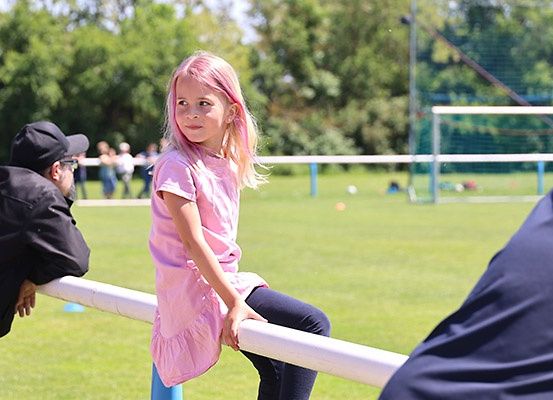Ein junges Mädchen mit rosa Haaren sitzt auf einem weißen Pfosten auf einem Sportplatz. Im Hintergrund beobachtet ein verschwommener Mann.