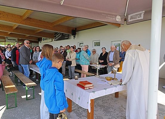 Ein Priester führt eine Zeremonie in einem Außenbereich durch, bei der ein mit einem weißen Tuch bedeckter Tisch und Bücher im Mittelpunkt stehen. Eine Menschenmenge steht hinter Bänken und beobachtet. Die Umgebung scheint ein religiöses Ereignis zu sein.