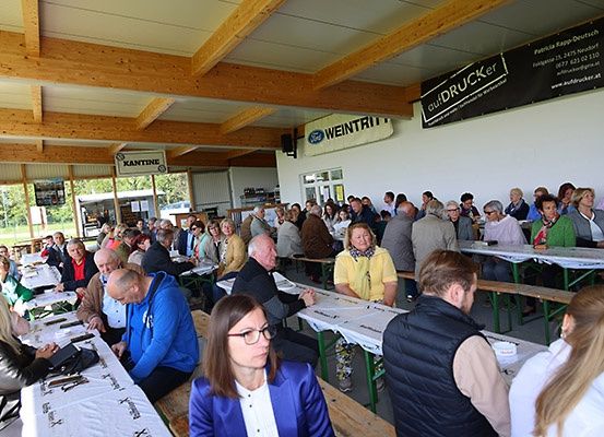 Eine große Gruppe von Menschen sitzt an langen Tischen in einem Pavillon. Die Umgebung ist im Freien, mit sichtbaren Holzbalken an der Decke. Einige Personen tragen Brillen und Jacken. An den Wänden hängen Banner, eines davon mit der Aufschrift 'Weintritt'.