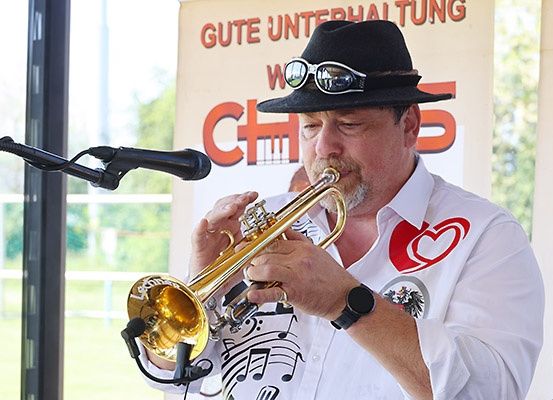 Ein Mann mit Hut und Sonnenbrille spielt Trompete bei einer Outdoor-Veranstaltung vor einem Mikrofon.